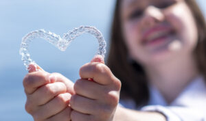 Smiling girl holding heart-shaped aligners. Plastic retainers for teeth alignment.